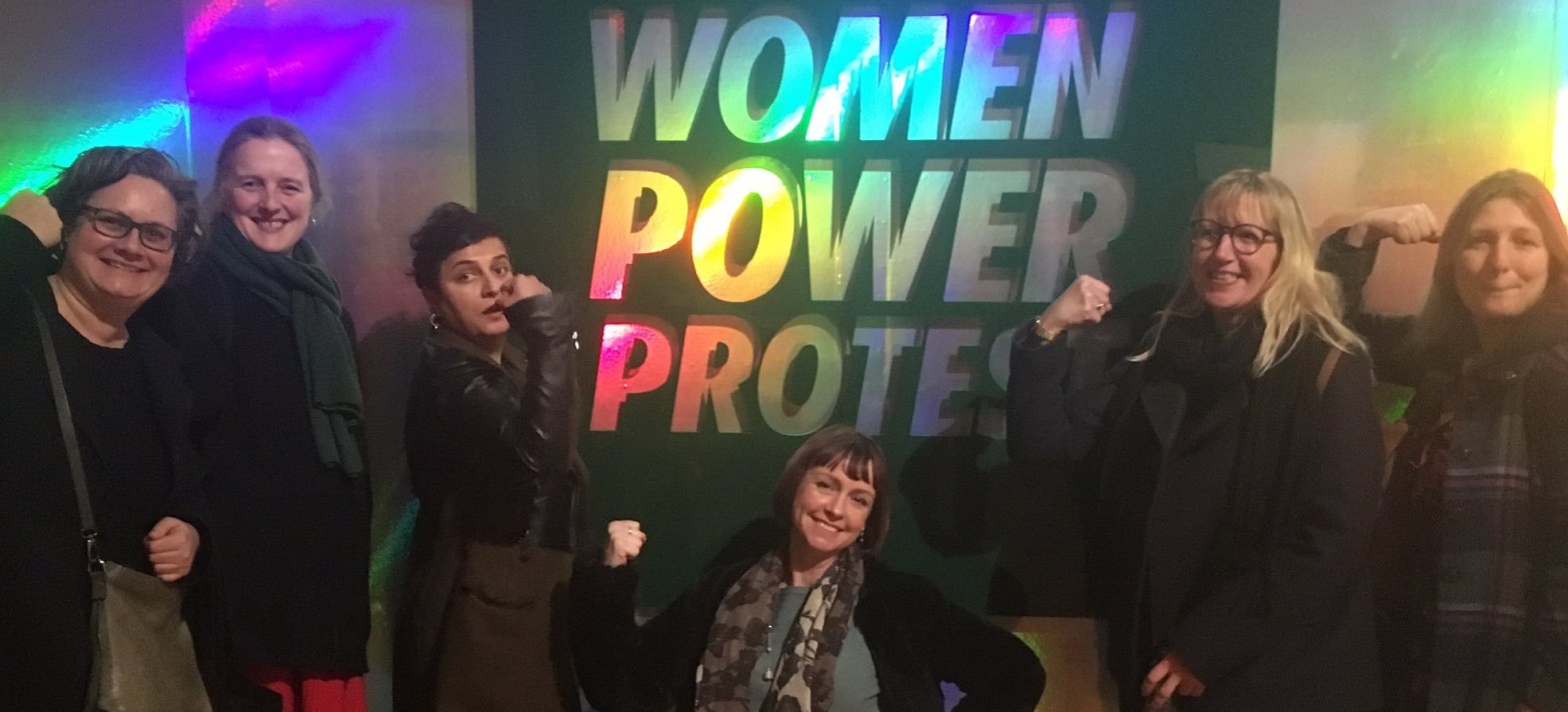 Space Invaders steering committee pose with power arms in front of the Women Power Protest holographic sign