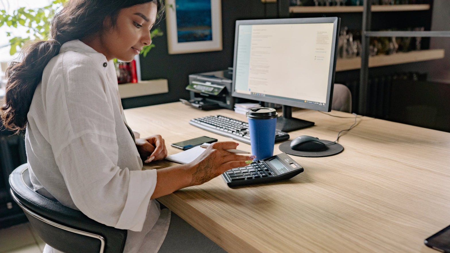 Woman sits at desk with a coffee using a large calculator